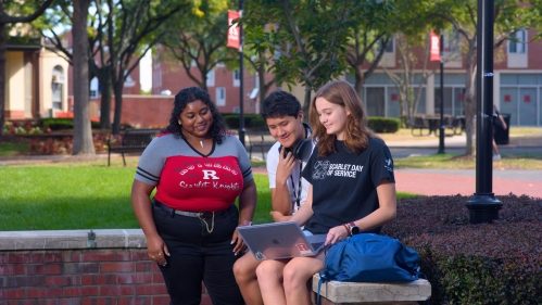 Three students smiling while working together on a laptop outside on campus