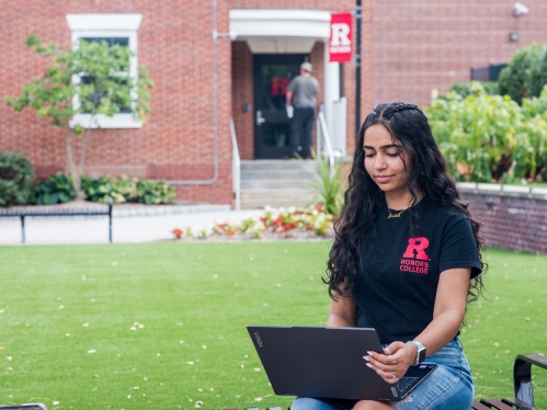 Student sitting on a bench using their laptop on campus during summer